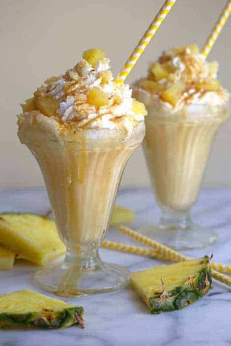 Two pineapple upside down milkshakes with yellow and white straws in them and pineapple wedges and straws decorated around them