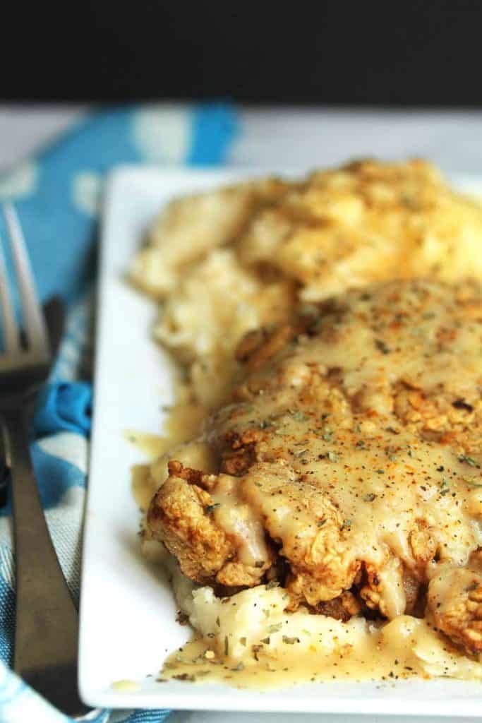 Chicken Fried Steak placed on a white plate and sitting on a bed of mashed potatoes, topped with gravy.