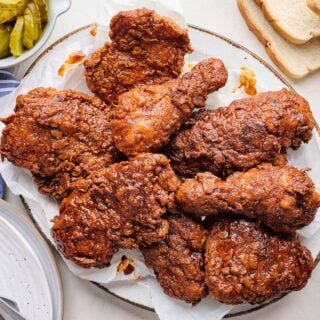 A platter of Nashville hot chicken on the table with a little bowl of pickles and slices of white bread.