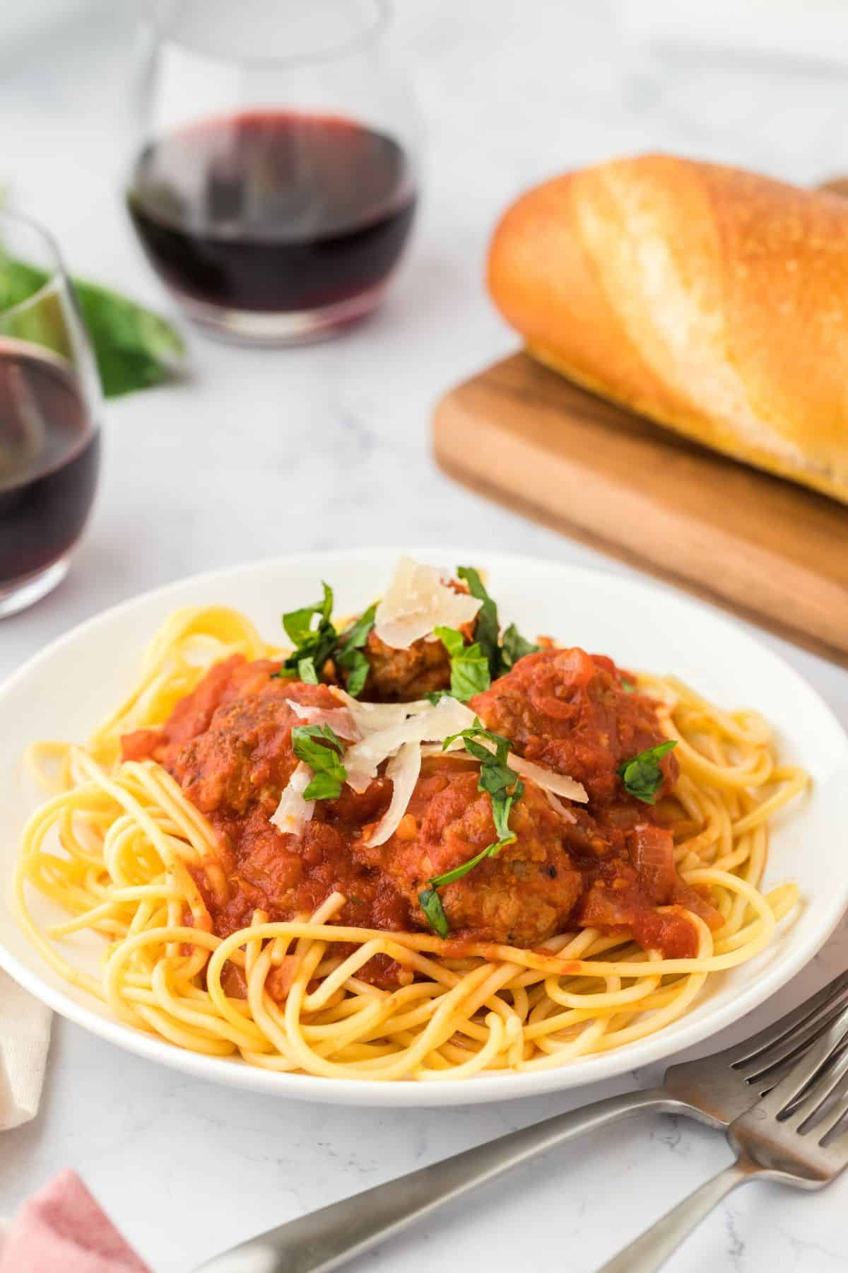 A plate of soul food spaghetti and meatballs on the table with a sprig of basil and loaf of bread in the background.