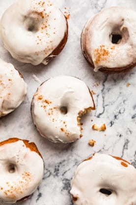 glazed pumpkin donuts against a white marble background with one in the center biten into