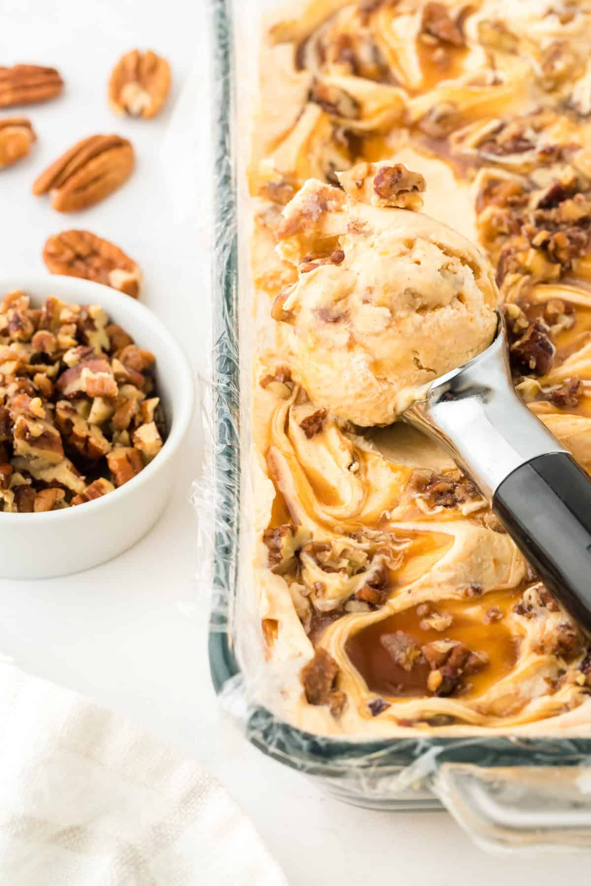 Sweet potato ice cream being scooped out to serve on white countertop with buttered pecans