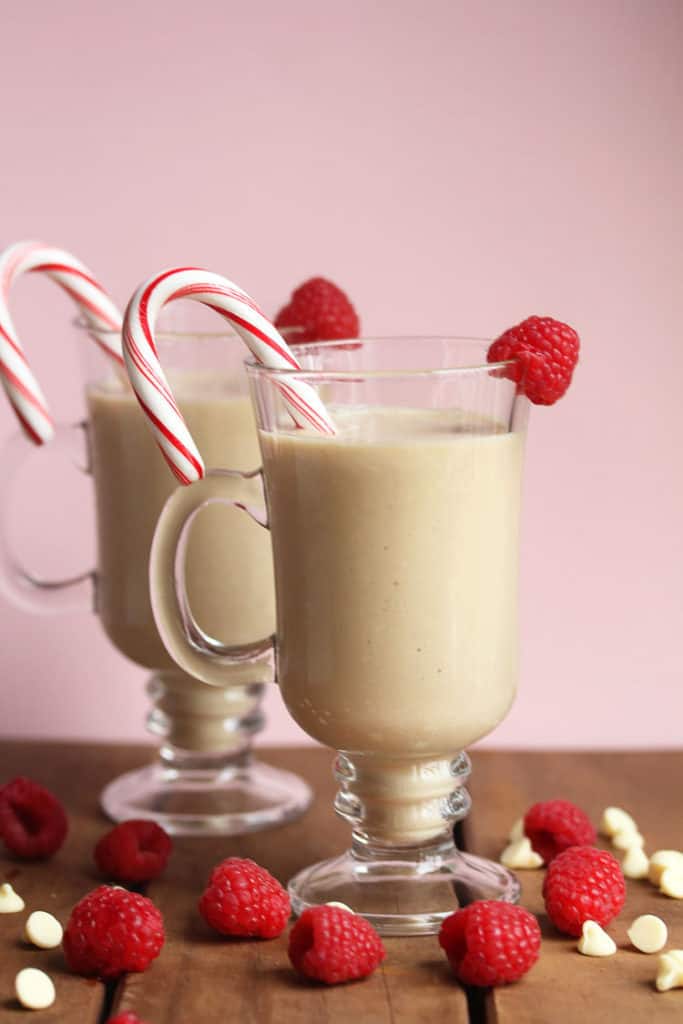 With fresh raspberries and white chocolate chips scattered around, two glass mugs filled with white chocolate mudslides, a raspberry and a candy cane as a garnish