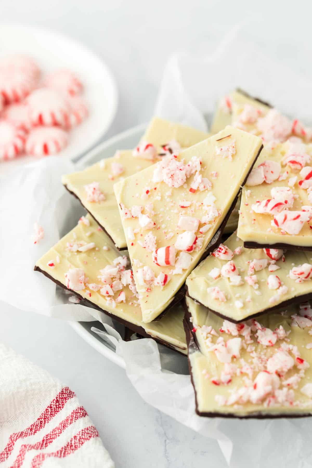 A pile of peppermint bark on a plate with a bowl of peppermint candy in the background.