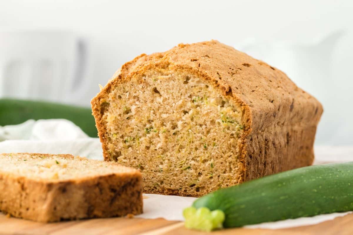 A loaf of zucchini bread on the table with a slice laying cut on the table and a whole zucchini to the side.