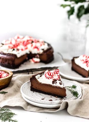 A slice of chocolate cheesecake topped with peppermint on white plate with full cheesecake behind