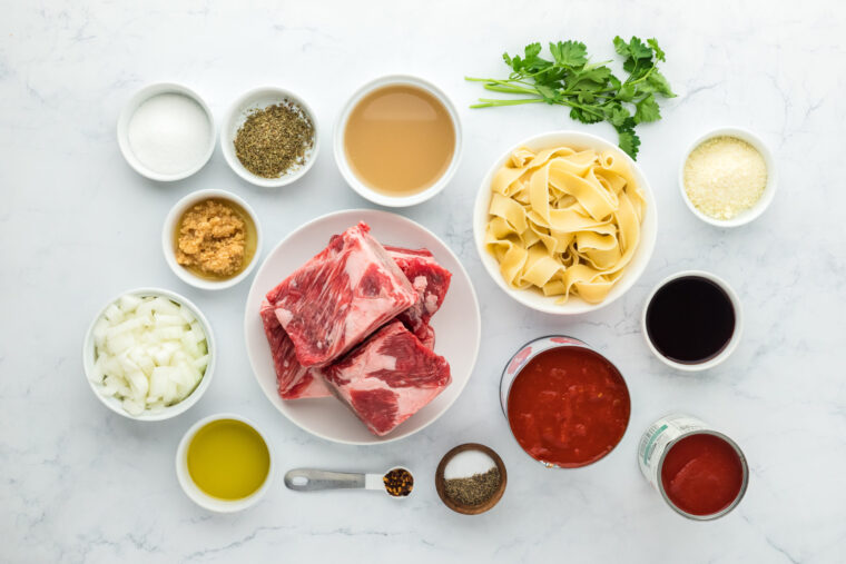 Short ribs, tomato sauce, herbs, spices in white bowls to make recipe on white countertop