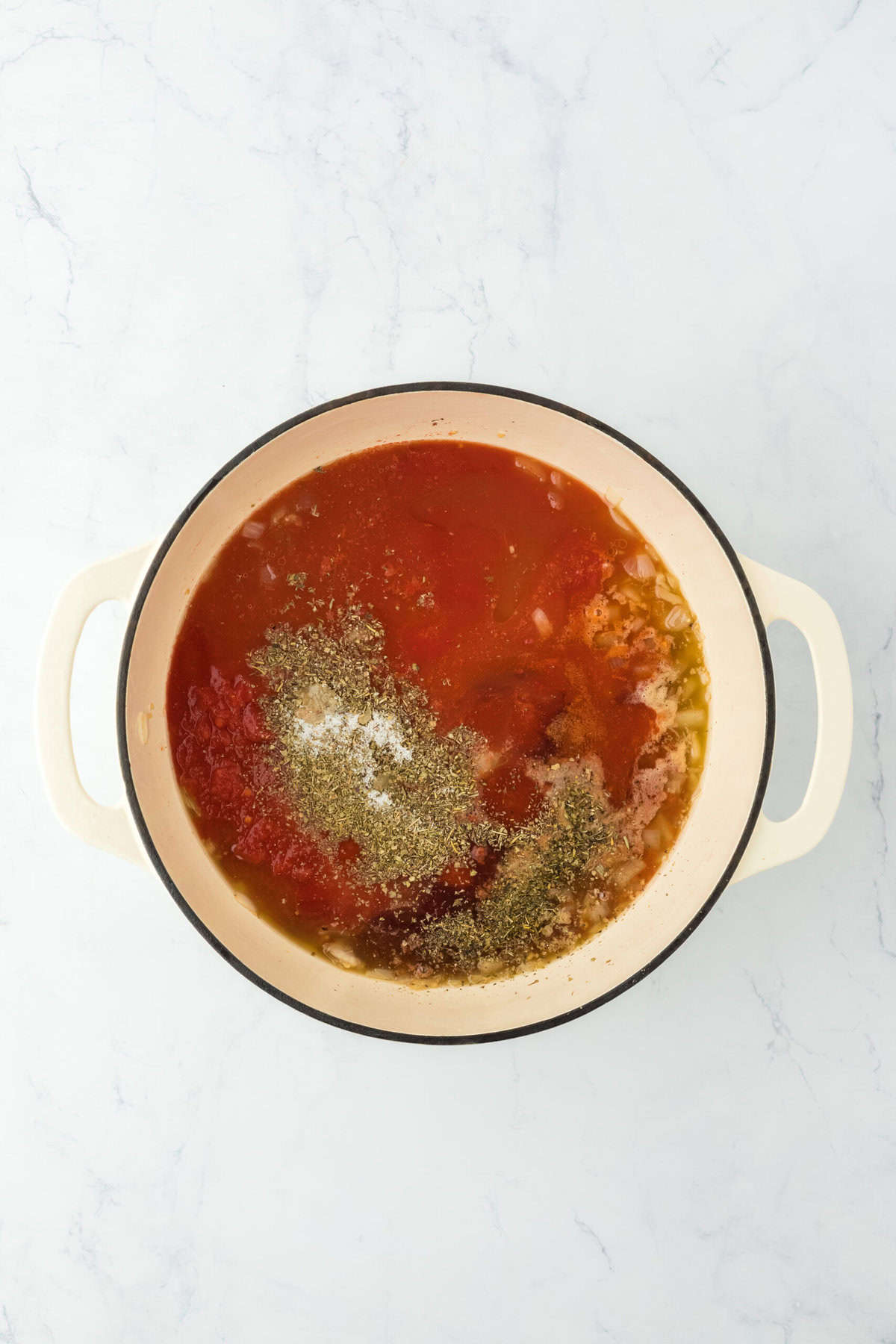 Tomato sauces and spices added to a dutch oven on white countertop