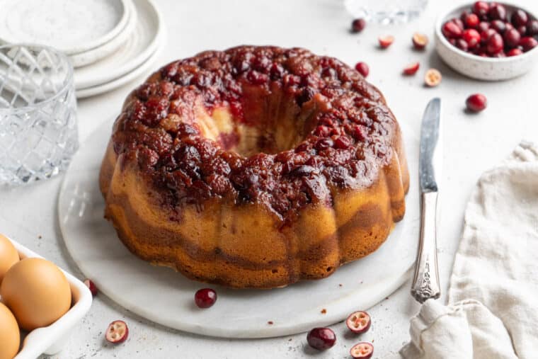 A cranberry bundt cake on a white platter on the table with a knife to the side and plates stacked in the background.