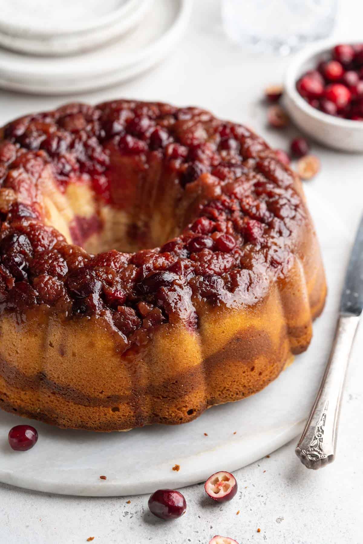 A cranberry pound cake on a platter with a little bowl of cranberries and stack of plates to the side.