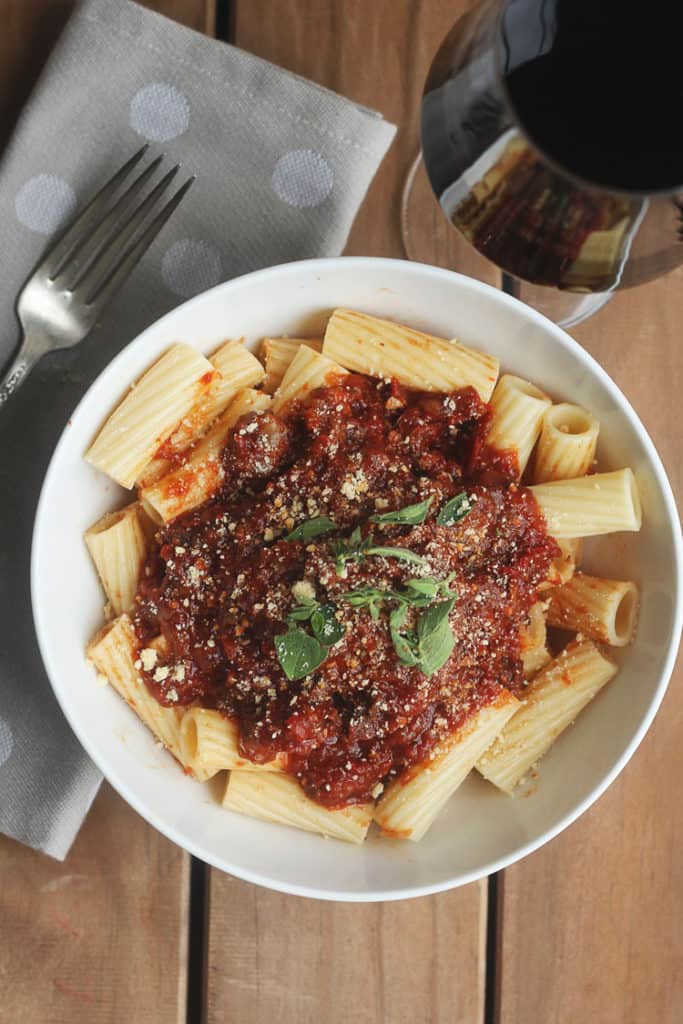 A bowl of delicious Italian pot roast with Bolognese Sauce over rigatoni with a glass of wine topped with basil and parmesan cheese
