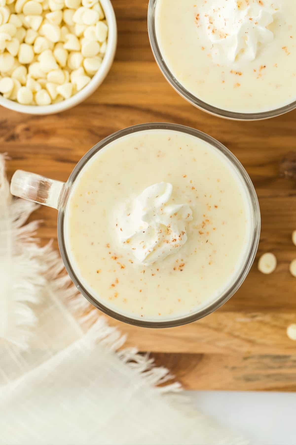A mug of white hot chocolate topped with whipped cream surrounded by another mug and a bowl of white chocolate chips.