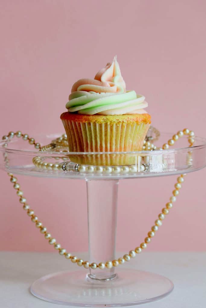 Pink and Green Vanilla Cupcake displayed on a clear plastic stand with a pearl necklace