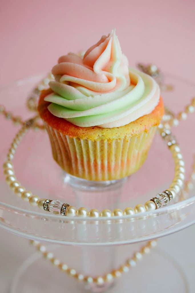 Pink and Green Vanilla Cupcake displayed on a clear plastic stand with a pearl necklace