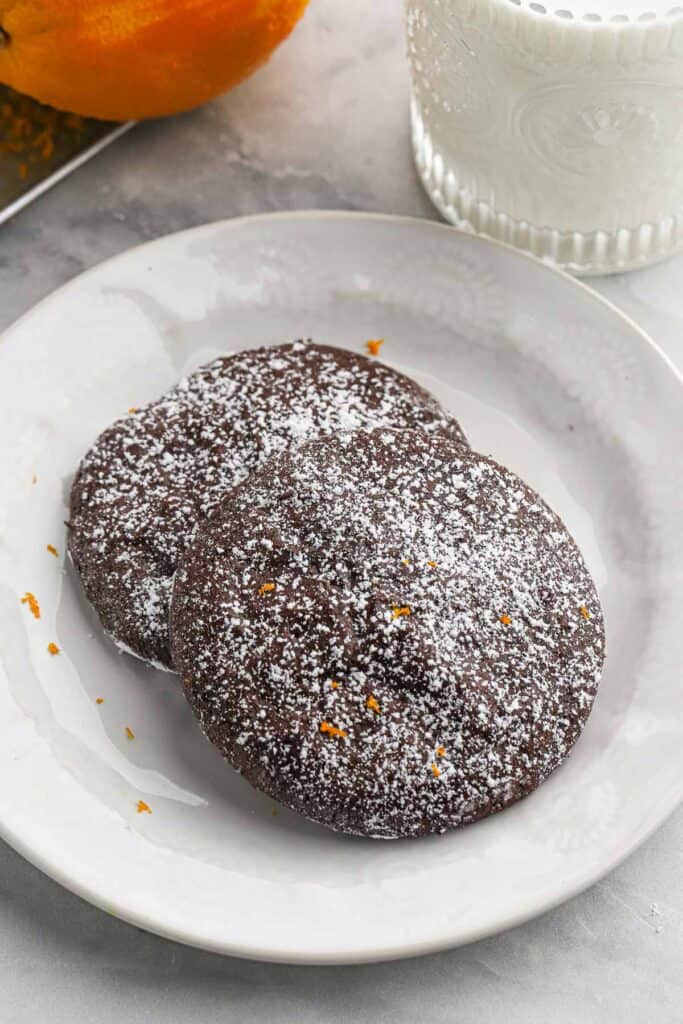 Overhead shot of a stack of Flourless Chocolate Orange Cookies topped with powdered sugar displayed on a white plate with oranges slices surrounding it