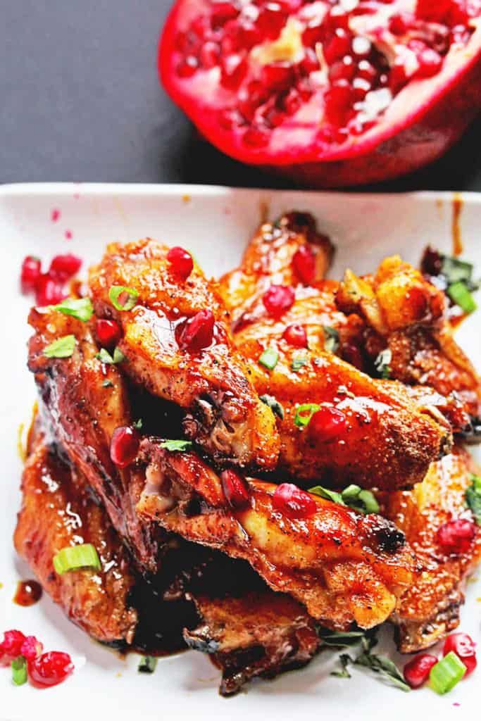 A close up of a pile of honey wings on a white plate sprinkled with pomegranate seeds