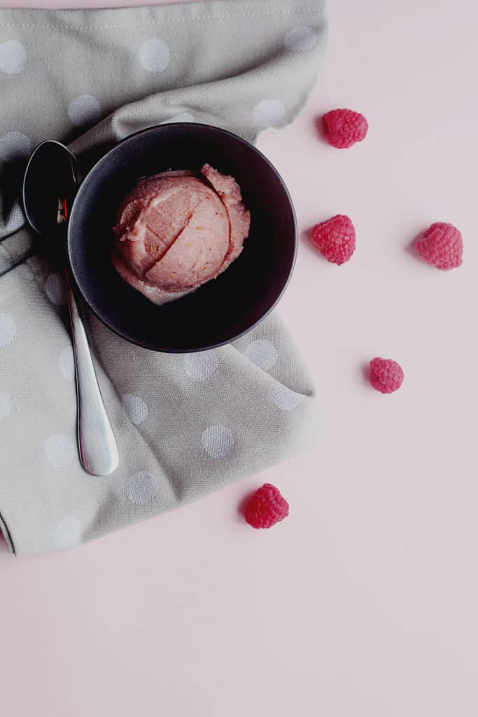 Overhead shot of one scoop of raspberry banana ice cream contained in a small bowl next to a spoon and fresh raspberries