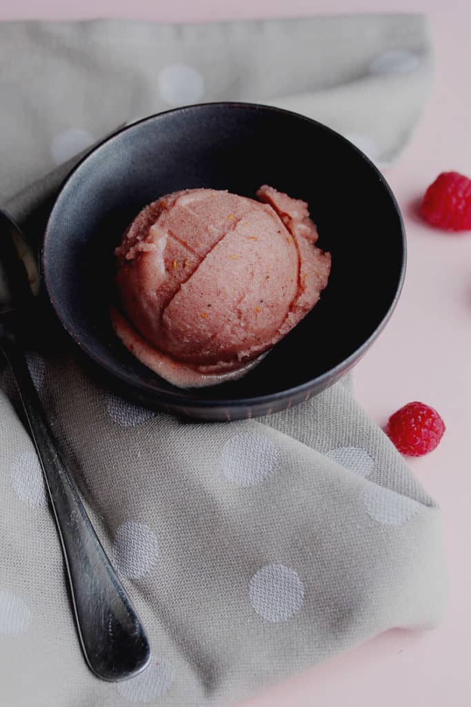One scoop of raspberry banana nice cream contained in a small bowl next to a spoon and fresh raspberries