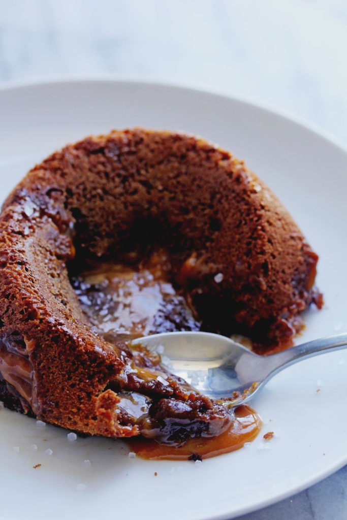 Close up of a Salted Caramel Lava Cake placed on a circular, white plate and opened by a spoon with caramel oozing out of the center