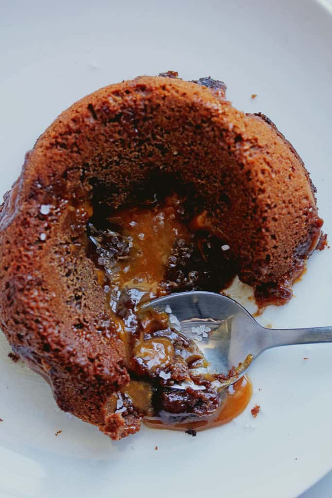 Overhead shot of a Salted Caramel Lava Cake placed on a circular, white plate and opened by a spoon with caramel oozing out of the center