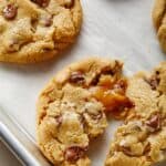 Salted caramel chocolate chip cookies on a baking pan with one broken in half with the caramel oozing out.