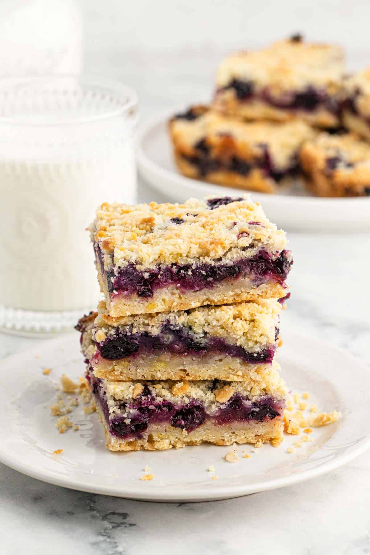 A stack of blueberry crumble bars on a plate with a glass of milk behind and a platter of more bars.