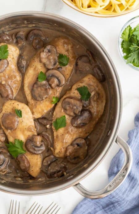 Closeup overhead shot of chicken marsala recipe with mushrooms and parsley in a pan, with fresh pasta and parsley nearby