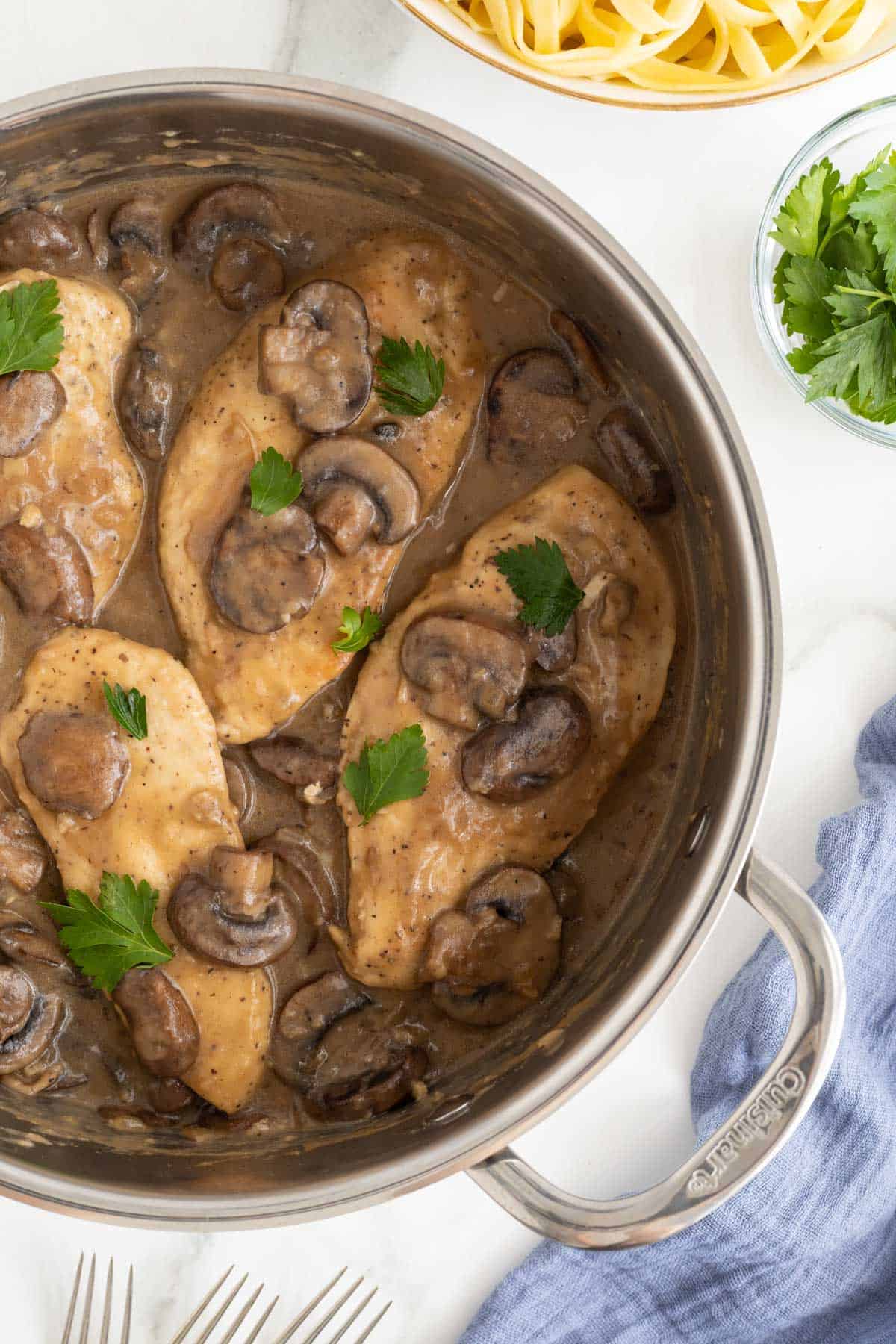 Closeup overhead shot of chicken marsala recipe with mushrooms and parsley in a pan, with fresh pasta and parsley nearby