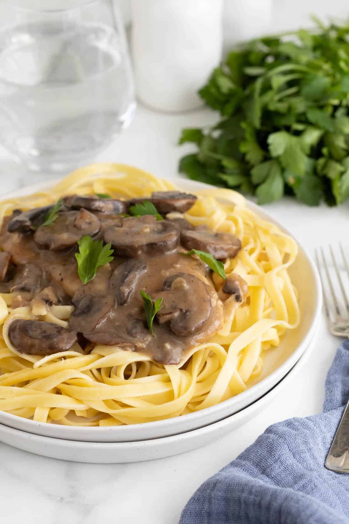 Plated serving of chicken marsala recipe over pasta, with fresh herbs and glass of water in the background