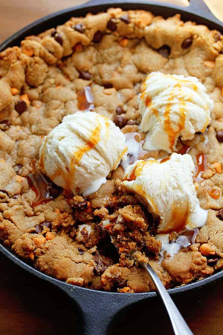 Overhead shot of a chocolate chip cookie cake with butterscotch chips served in a black cast iron skillet and topped three scoops of vanilla ice cream and a spoon in it