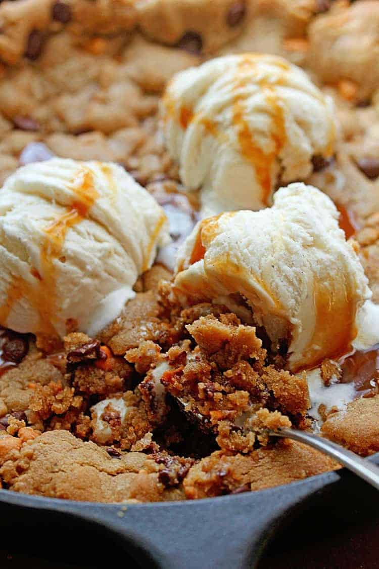 Close up of a chocolate chip cookie cake with butterscotch chips served in a black cast iron skillet and topped three scoops of vanilla ice cream and a spoon in it