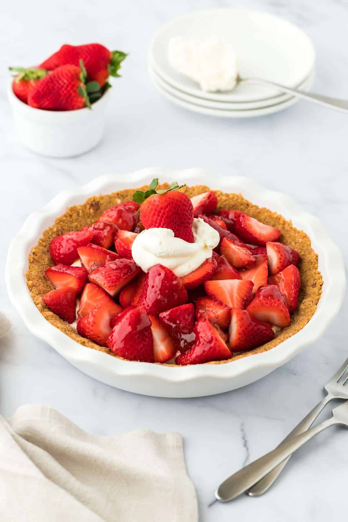Strawberry custard pie on a table with a bowl of strawberries and a stack of plates in the background.