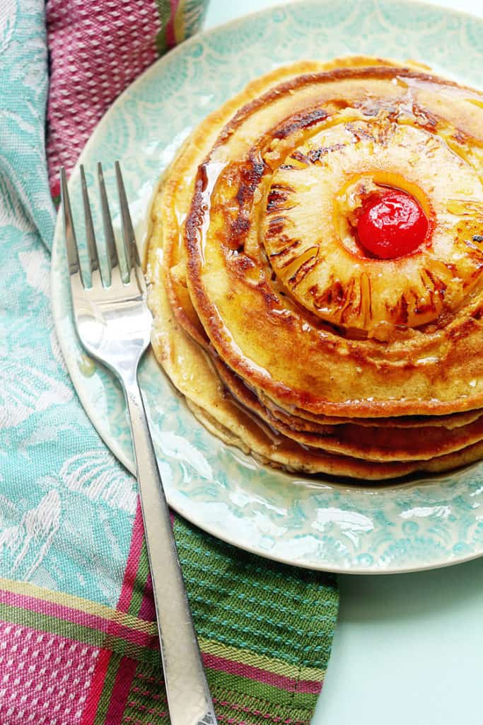 Overhead shot of a stack of pineapple upside down pancakes topped with baked pineapple and cherry in the center served on a light blue plate with a fork