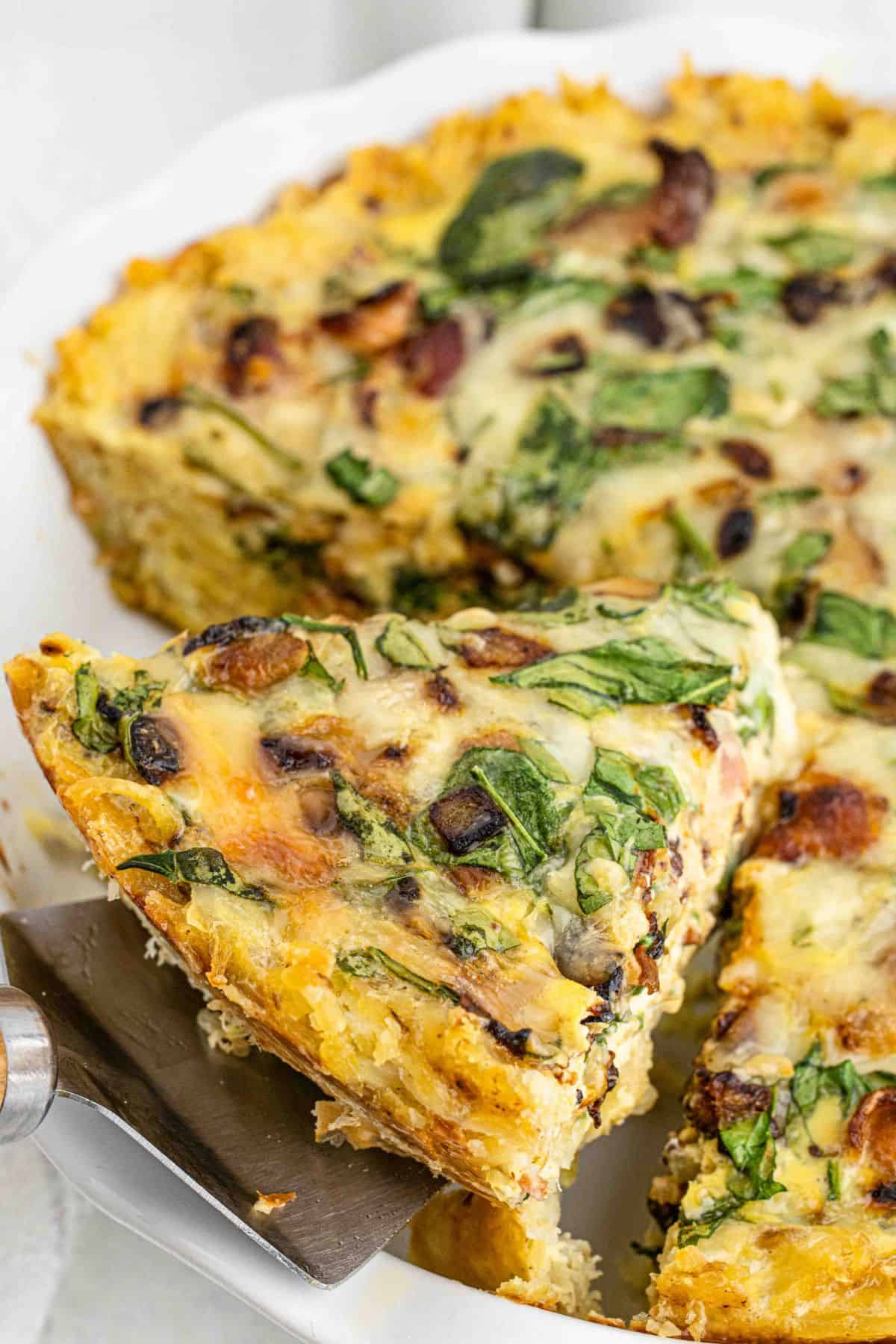 Spinach bacon hash brown quiche in a pie plate with a spatula lifting up a slice.