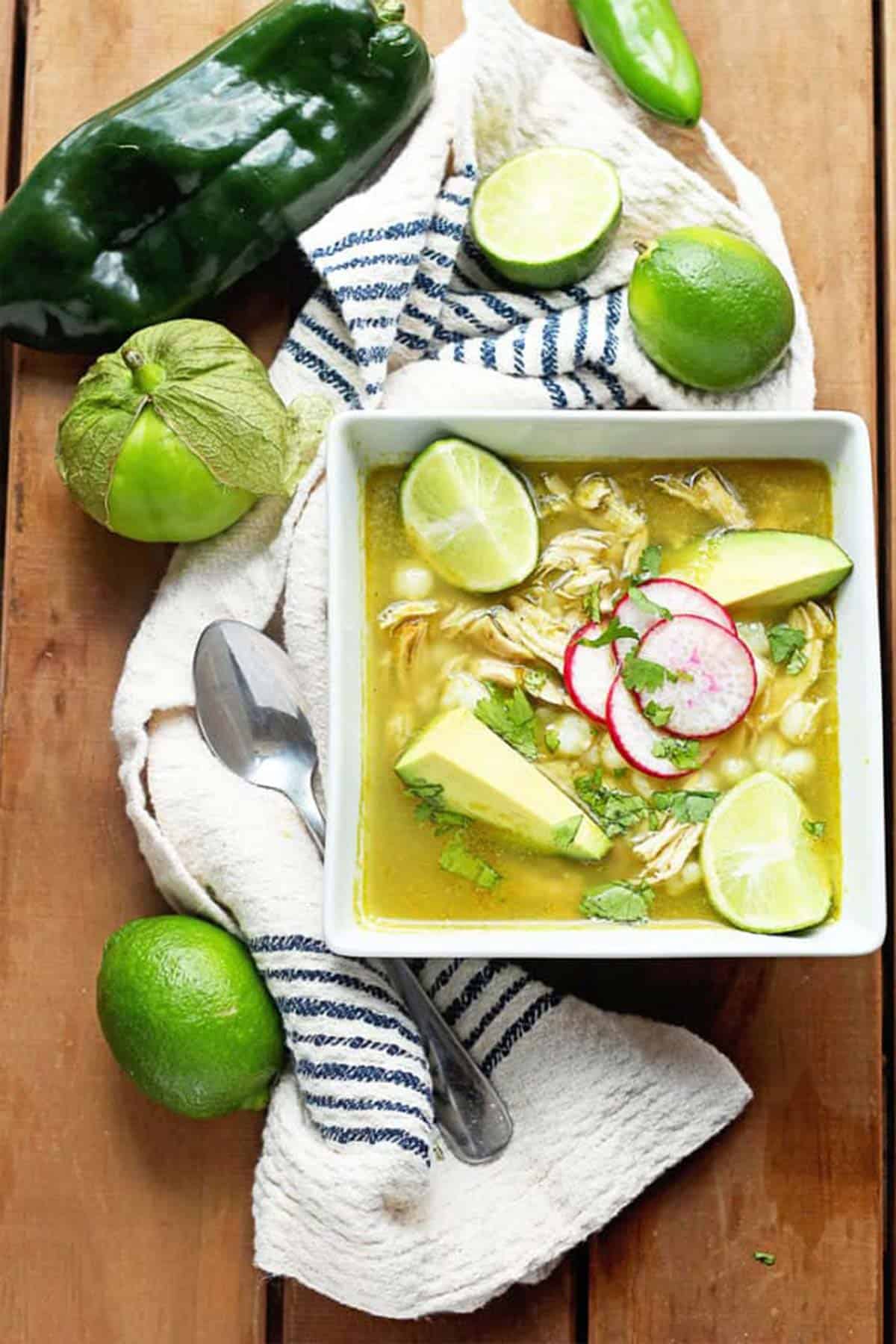 A bowl of verde pozole with chicken on the table with limes, tomatillos and chile.