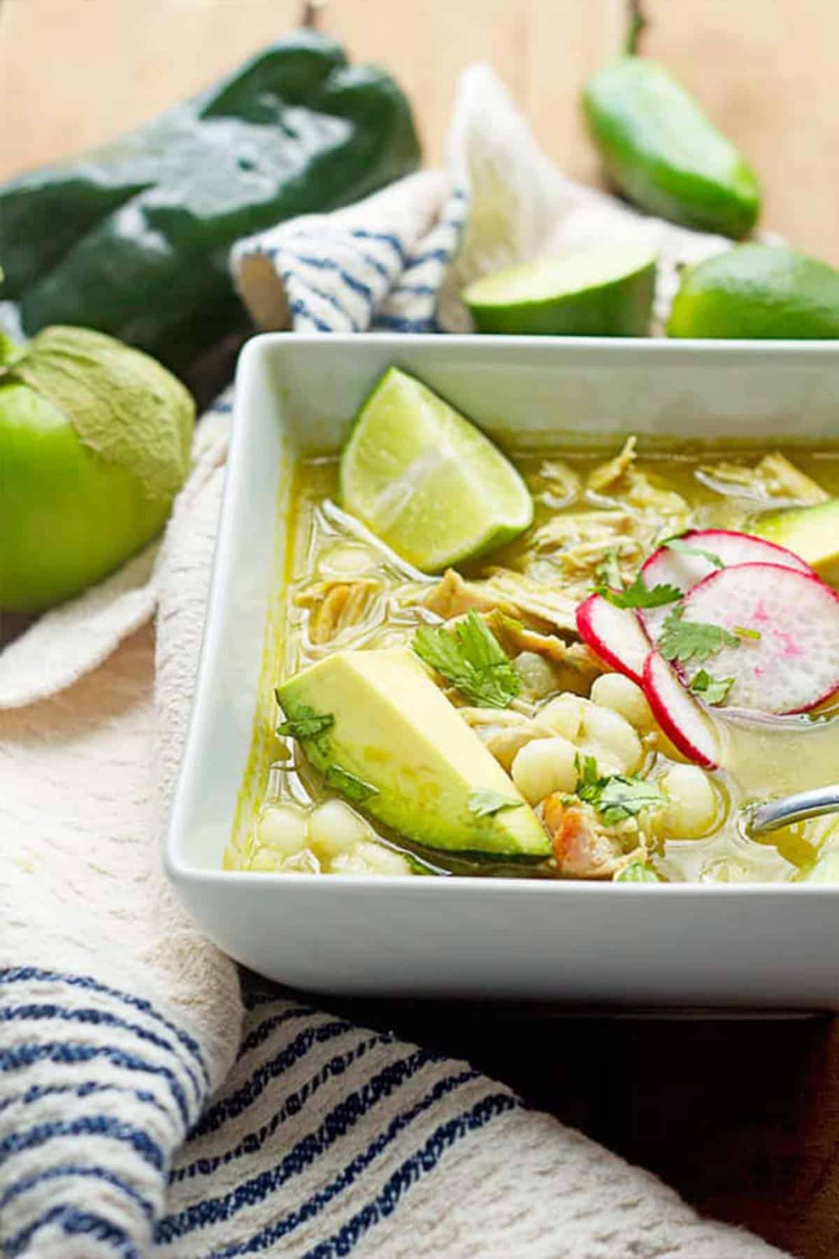  A bowl of Chicken Posole Soup in a large white bowl garnished with avocado, radishes and limes with a spoon in the bowl.