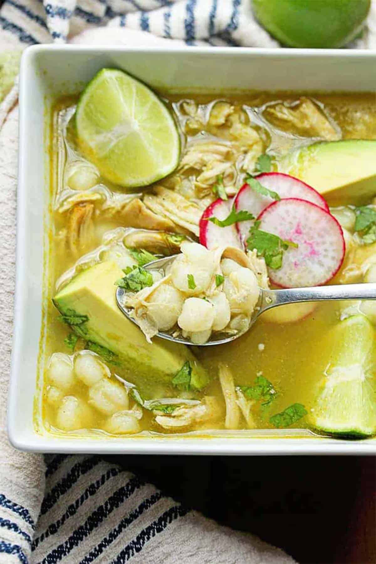 A bowl of chicken posole verde with a spoon lifting a some from the bowl.