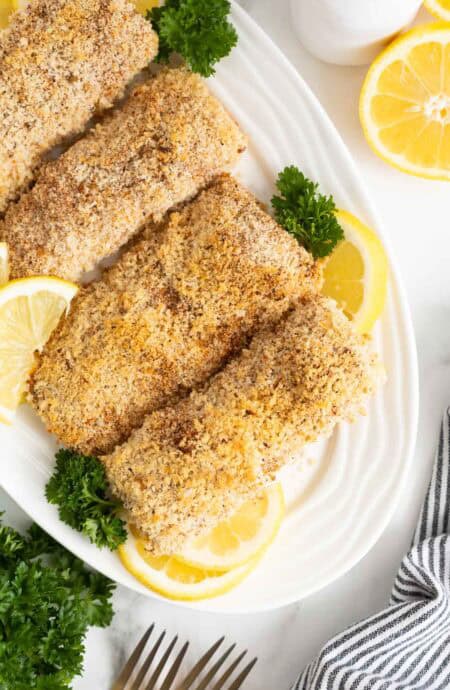 Overhead shot of several lemon panko crusted fish fillets on a white serving platter, garnished with parsley and lemon slices