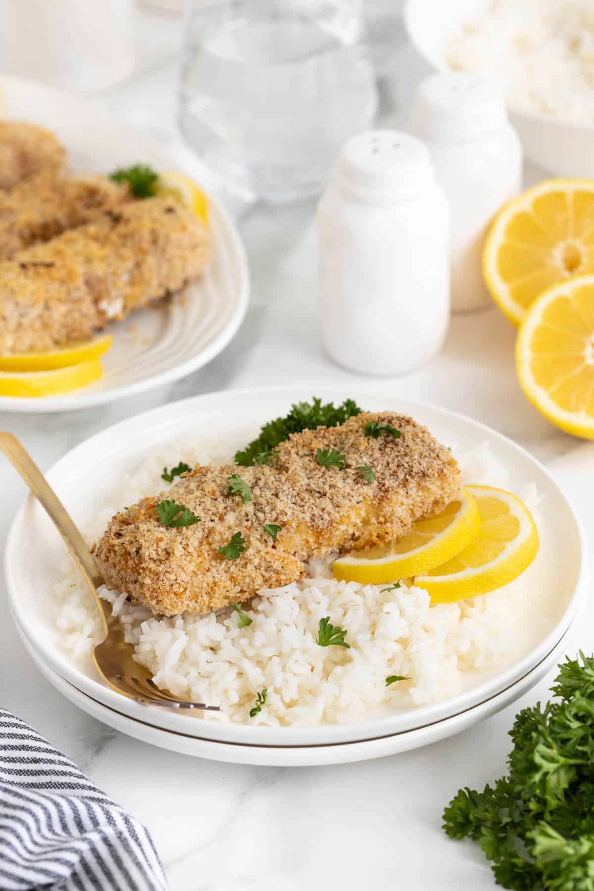 Lemon panko crusted fish served over rice, garnished with parsley and lemon slices, with salt shakers, lemon slices, and a platter in the background