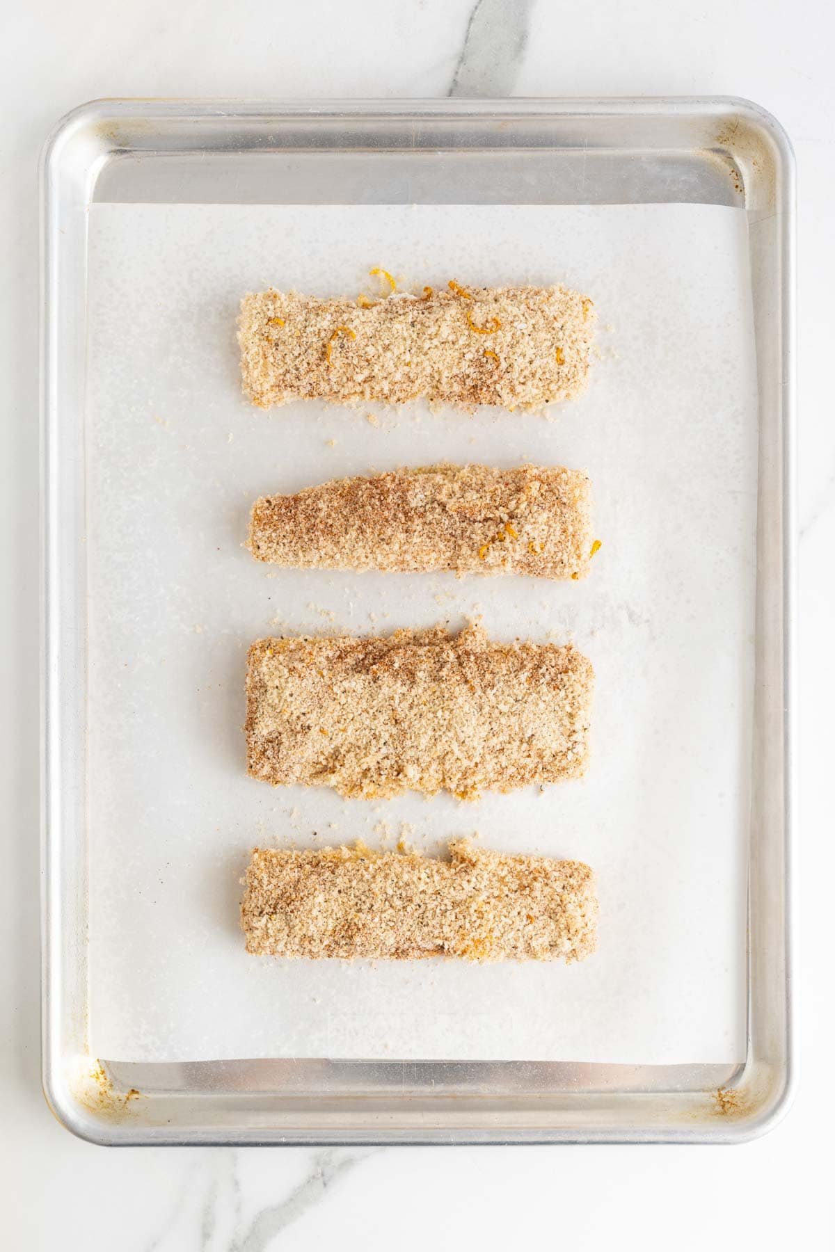 Four breaded fish fillets arranged on a parchment-lined baking sheet, ready for the oven