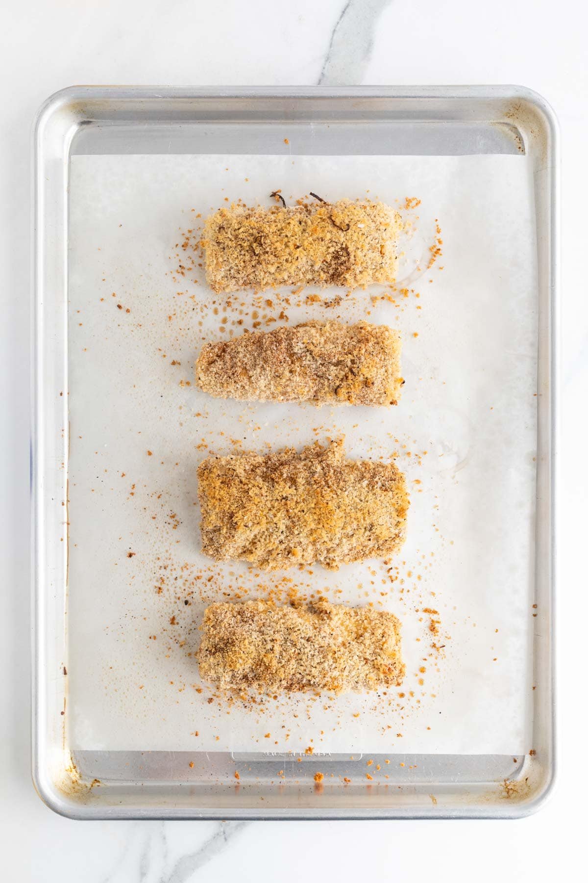 Baked breaded fish fillets on a parchment-lined baking sheet with golden panko crumbs scattered around