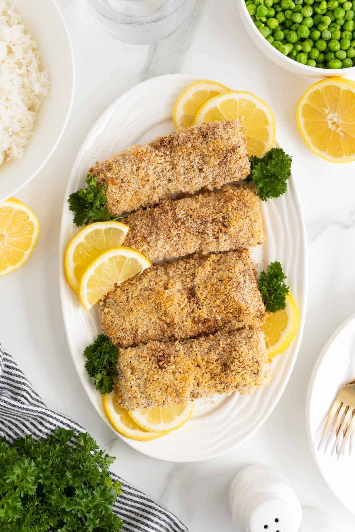 Cooked lemon panko crusted fish fillets served on a white platter, garnished with lemon slices and parsley, with bowls of rice and peas nearby