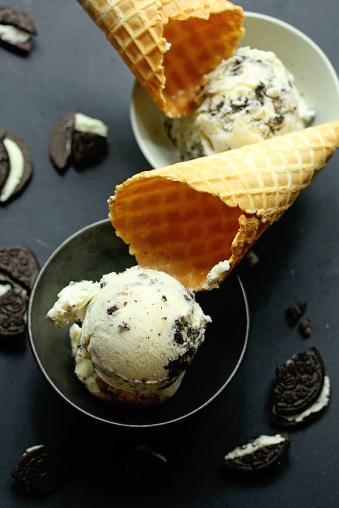 A close up of cookies and cream ice cream served in two bowls, one white and the other black, with waffle cones and pieces of oreo cookies next to them