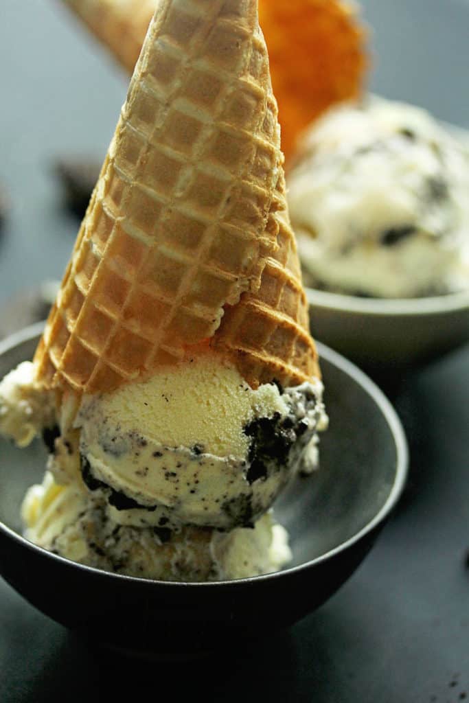 Close up of a scoop of Cookies and Cream Ice Cream served in a black bowl with a waffle cone sitting on top of it and another bowl of it in the background