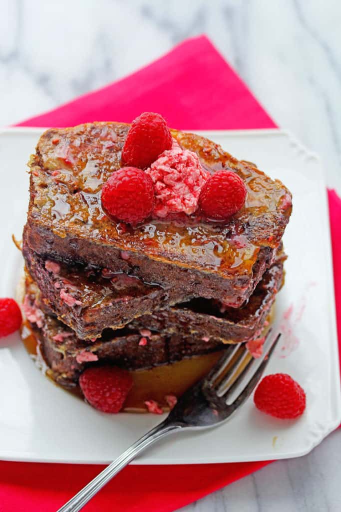 Overhead shot of a stack of Banana Bread Cinnamon Swirl French Toast served on a square, white plate and topped with fresh raspberries and syrup