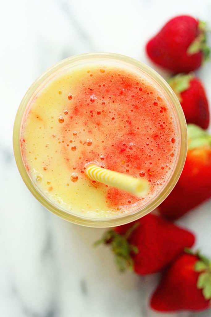Overhead shot of a tropical smoothie recipe with a yellow and white striped straw in it and fresh strawberries next to it