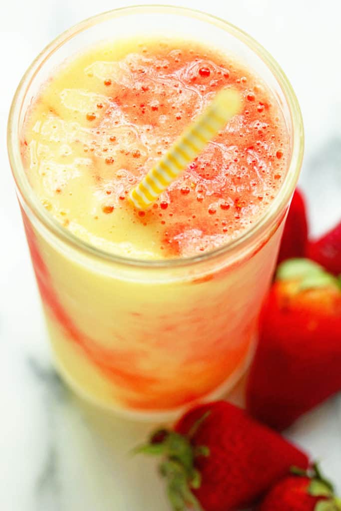 Close up of a tropical smoothie with a yellow and white striped straw in it and fresh strawberries next to it