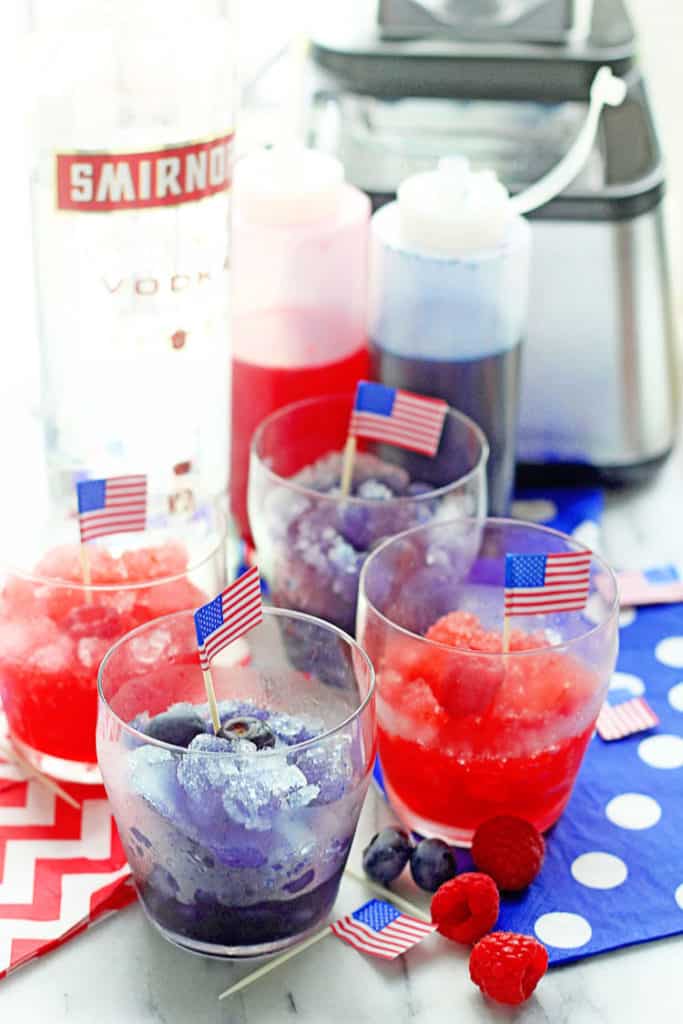 Red and blue spiked snow cones served in small clear glasses and a bottle of vodka and red and blue syrup in the background
