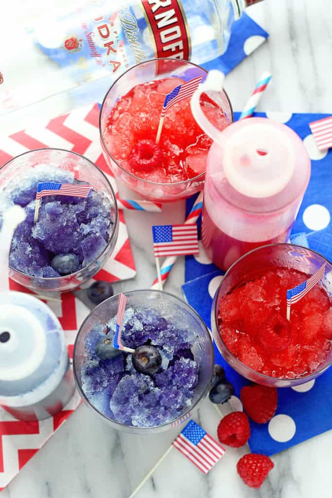 Overhead shot of red and blue spiked snow cones served in small clear glasses with USA flags in them and a bottle of vodka nearby