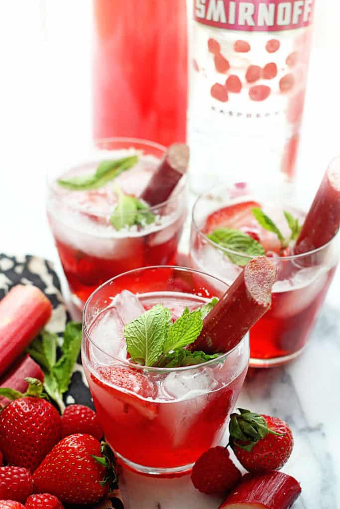 Three glasses of Berry Rhubarb Punch garnished with pieces of mint and rhubarb and a bottle of smirnoff vodka, fresh strawberries and rhubarb nearby